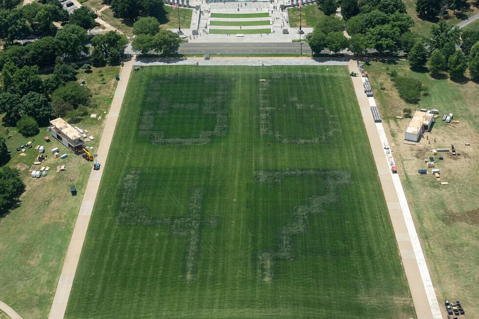 Aerial view of a large lawn with mowed patterns forming the numbers "84" and "47." Pathways border the grass on either side, and various construction materials and equipment are positioned around the area. Trees and a paved area with steps and flags are visible in the background.