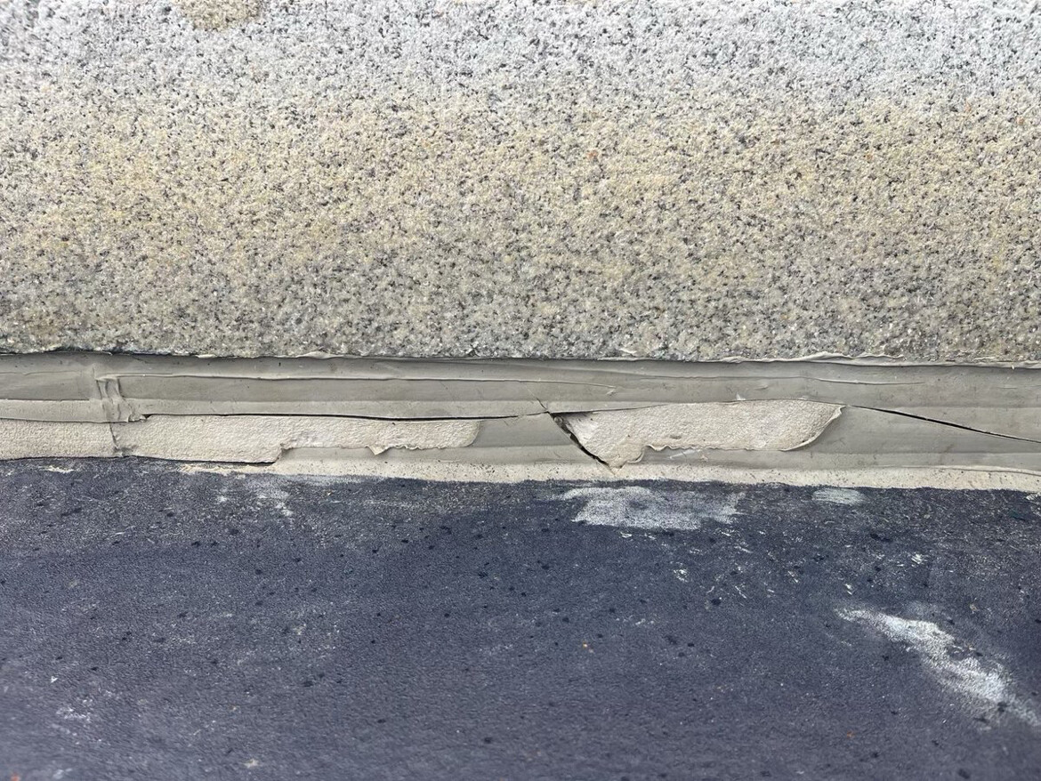 Close-up view of a textured wall meeting a floor surface, showcasing a line of caulking between them. The image highlights areas of peeling and cracking in the caulking, indicating wear and potential need for maintenance. The wall has a speckled appearance, while the floor below is darker and smooth, with visible signs of wear.