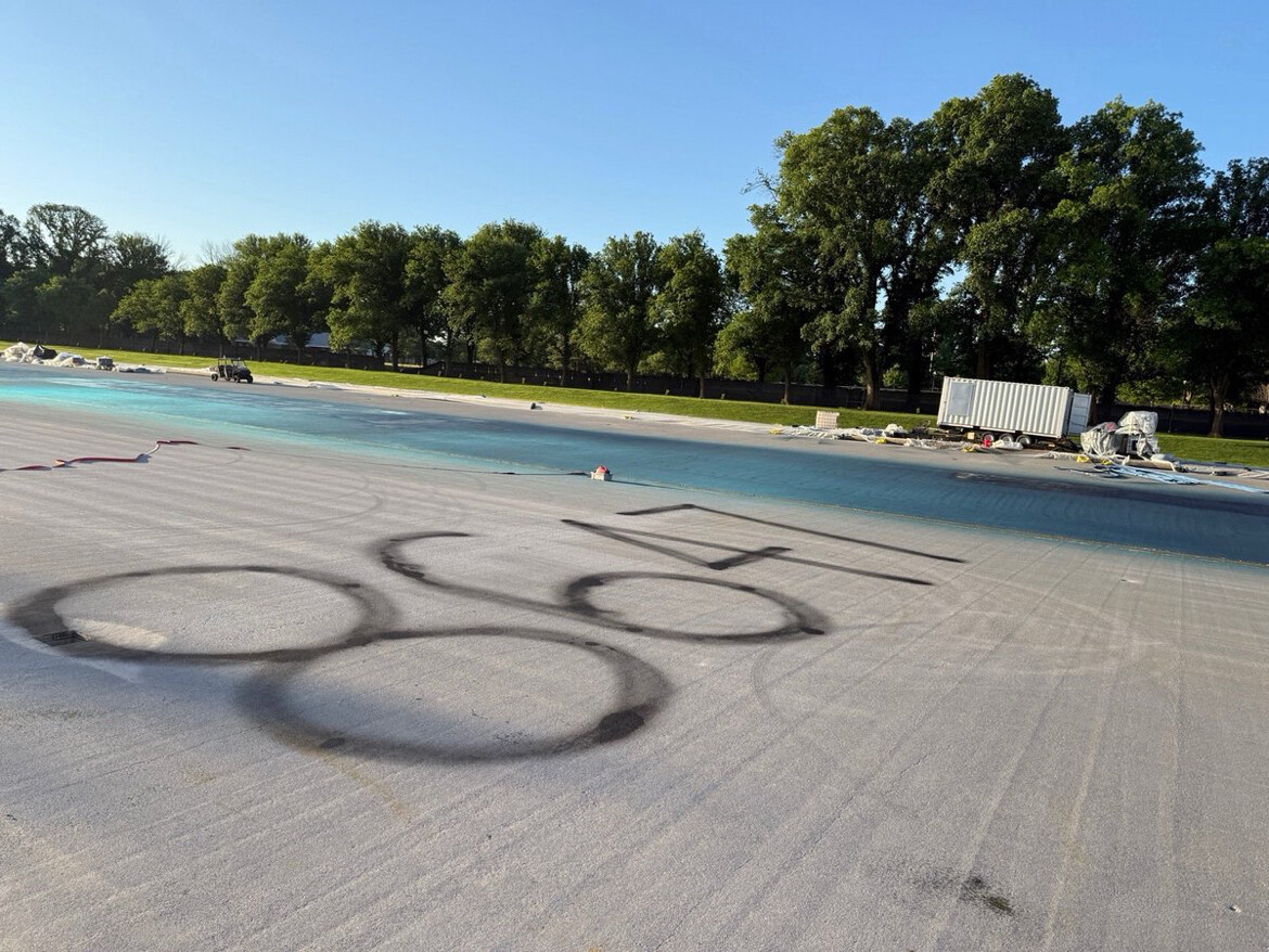 Empty pool area with large black stencil of the Olympic rings on the ground, surrounded by trees and construction equipment.