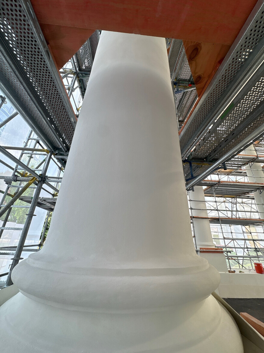 A large white architectural column under construction surrounded by scaffolding. The column is painted with a bright white finish, and the scaffolding is composed of metal frames and wooden planks. The setting appears to be inside a building with large windows and additional columns visible in the background.