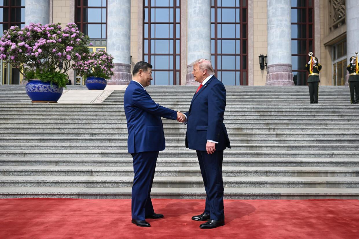 Two people are shaking hands on a red carpet in front of a large building with tall columns and steps. Both are dressed in formal dark suits, and there is a significant potted plant with pink flowers to the left. Two uniformed guards are visible in the background.