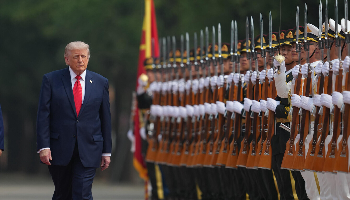 An older man in a navy suit with a red tie walks alongside a line of uniformed soldiers holding rifles with bayonets. The soldiers stand in a formation, wearing dark uniforms with white gloves, and have a flag visible in the background. The setting is outdoors with trees in the background.