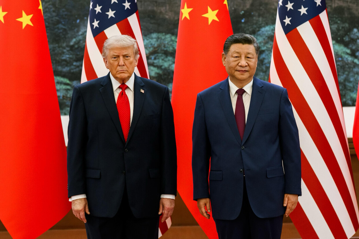 Two men in suits stand side by side in front of flags. The man on the left wears a navy suit with a red tie and the man on the right wears a dark suit with a dark tie. Behind them are two U.S. flags with red and white stripes and a blue field with white stars, and two Chinese flags with red fields and yellow stars.
