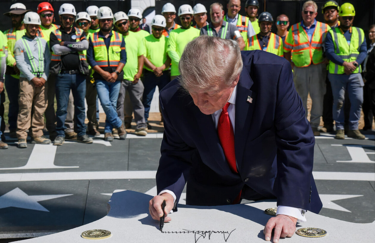 A man in a suit with a U.S. flag pin is signing a large stone slab with a marker. In the background, a group of construction workers wearing hard hats and high-visibility clothing are standing in a line, watching the signing. The setting appears to be outdoors with construction-related elements visible.