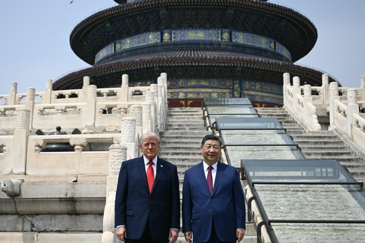 Two men in suits stand outdoors in front of a traditional Chinese temple with intricate designs and a sloping roof. The temple features ornate stone railings and stairs. The man on the left wears a dark suit with a red tie, and the man on the right wears a dark suit with a blue tie. The setting appears to be clear and during the daytime. A bird is visible flying above the temple.