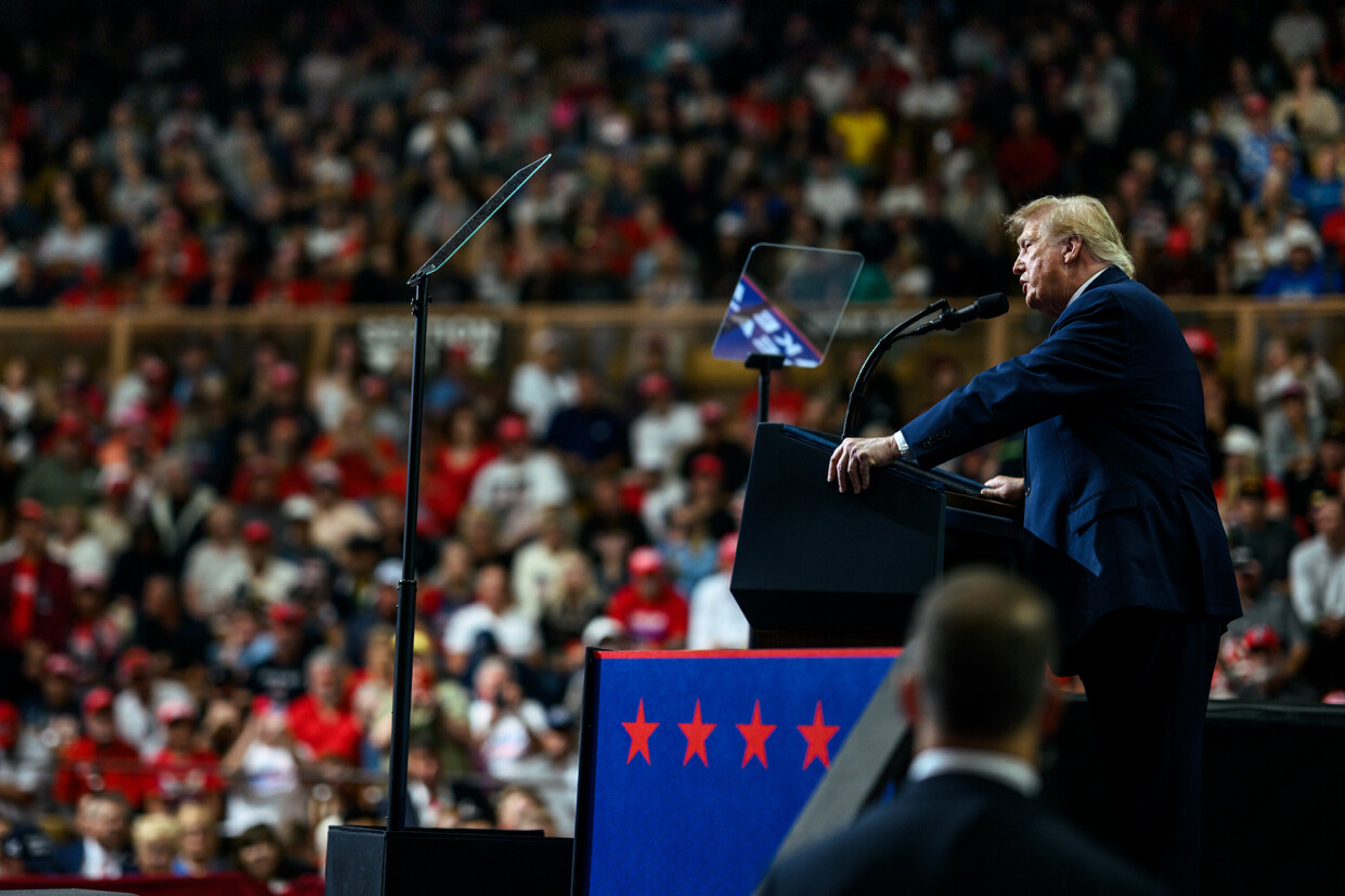 A person is speaking at a podium with teleprompters to a large audience in an indoor venue. The podium has a blue panel with five red stars. The crowd in the background is blurred, with many people dressed in red clothing. A security personnel is visible in the foreground.