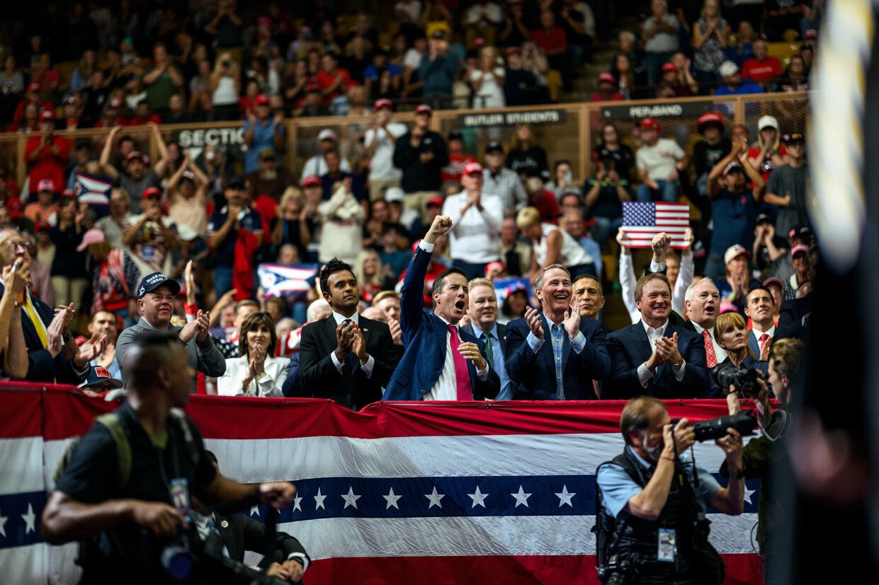 A lively crowd is gathered at an indoor event, featuring people dressed in formal attire, some wearing red caps. Attendees hold American and Puerto Rican flags, clapping and cheering behind a patriotic banner with stars and stripes. A photographer in the foreground captures the moment, while signs labeled "SECTION 215" and "PERIMETER" are visible above the spectators.
