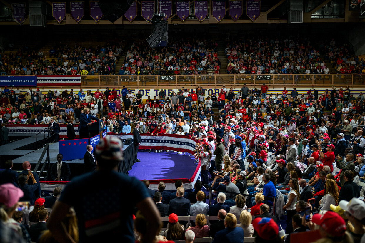 A crowded indoor rally with a stage features a person speaking at a podium, surrounded by multiple officials. The audience, wearing red hats and patriotic attire, fills the venue, and banners with phrases such as "MAKE AMERICA GREAT AGAIN!" are visible. The background displays sports championship banners for different events.