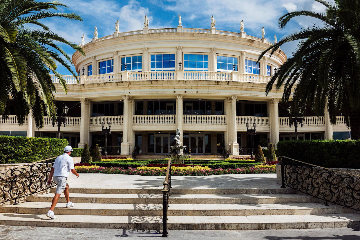An elegant, two-story neoclassical building with tall columns and large windows, surrounded by palm trees. A set of steps leads up to the entrance, with a decorative fountain in front. A person wearing light clothing and a cap is walking up the steps in the foreground.