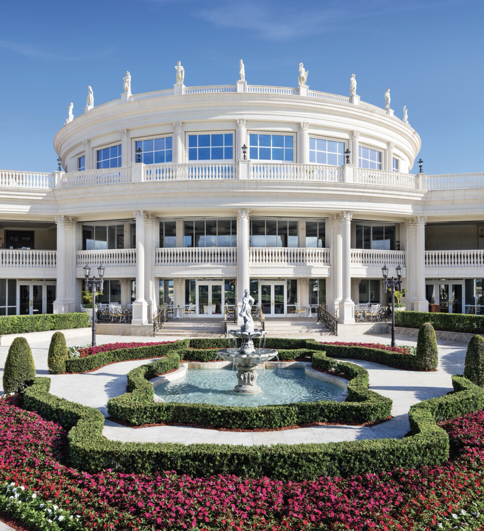 A grand, white circular building with classical architecture features multiple columns and statues on its rooftop. The façade includes large windows and two levels of balconies with balustrades. In the foreground, there is a landscaped garden with a central fountain, surrounded by neatly trimmed hedges and colorful flowers. The sky is clear and blue.