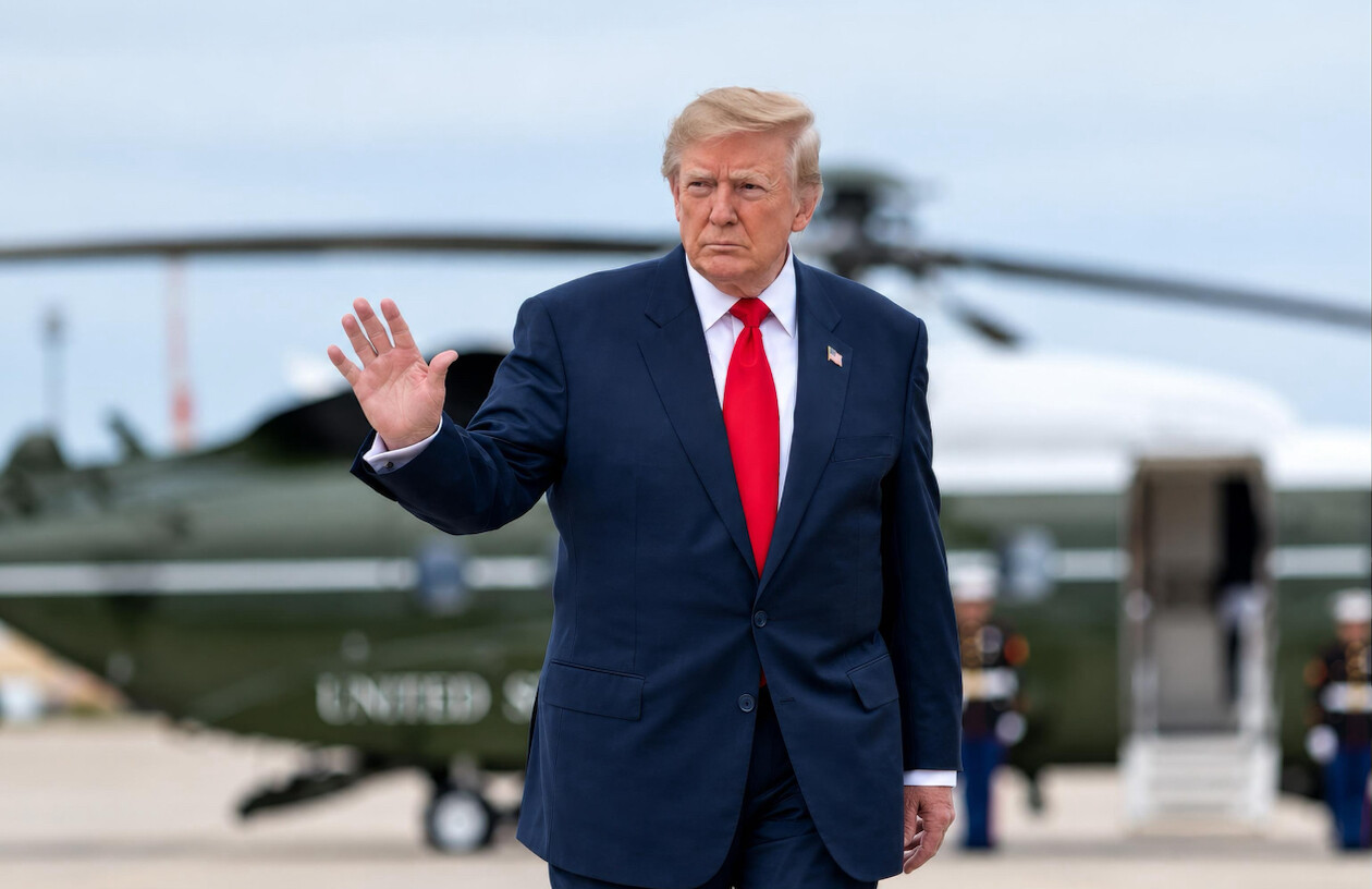 A person in a dark blue suit with a red tie is walking with one hand raised. The background features a helicopter on the ground, marked "UNITED STATES." The individual is outdoors and appears to be in a public setting, with people in uniform visible in the background.