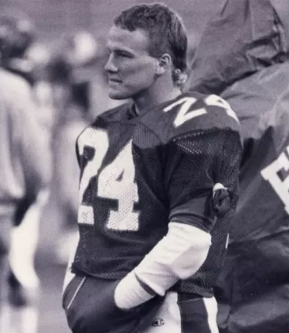 A black and white photograph of a person in a football uniform, wearing a jersey with the number 24. The individual is standing with hands in pockets, facing to the side. The background shows a blurred figure and another person partially visible, wearing a hooded jacket.