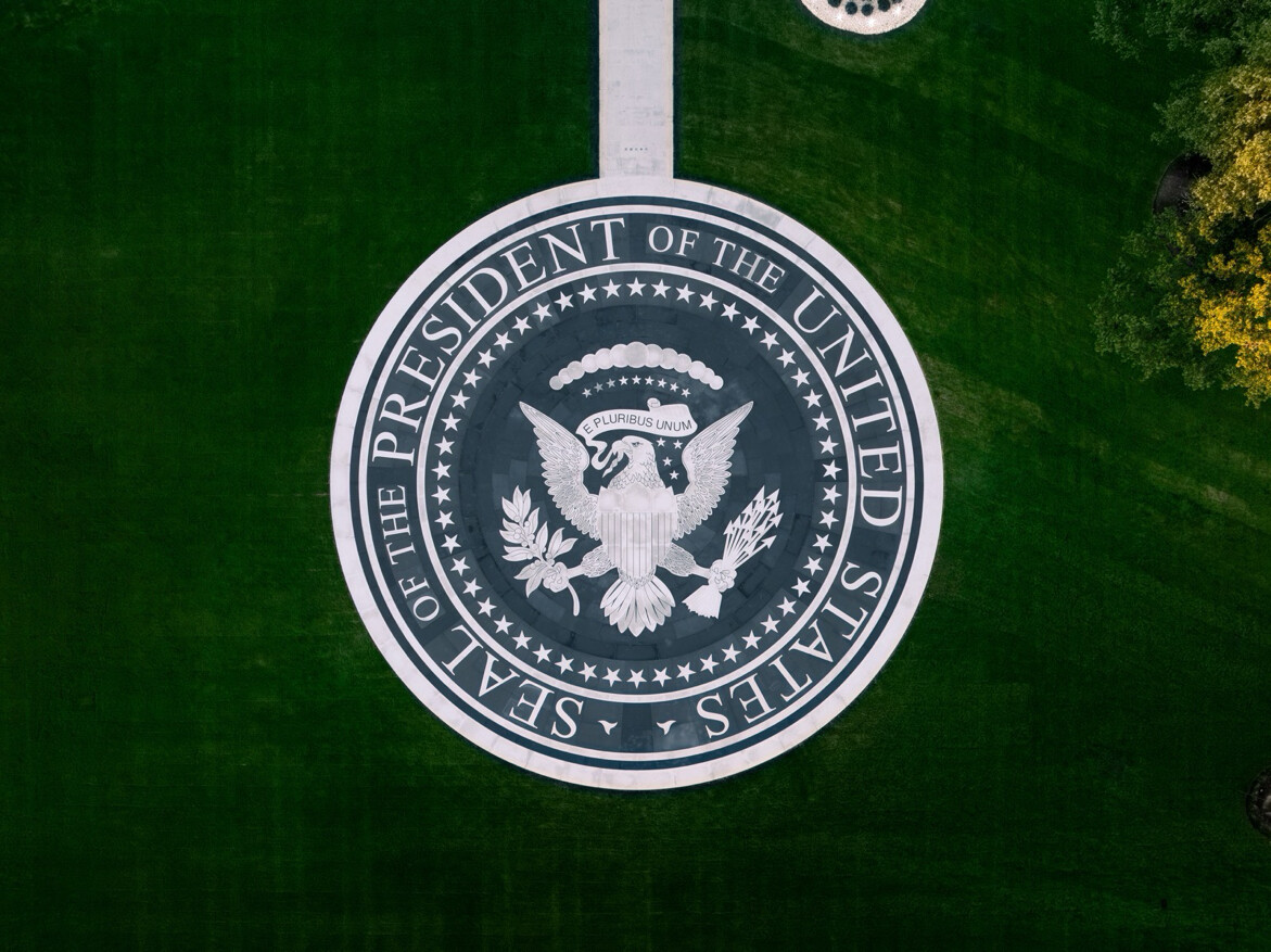 Aerial view of the Seal of the President of the United States on a circular pavement, surrounded by green grass. The seal features a bald eagle holding an olive branch and arrows, with a banner reading "E Pluribus Unum" above its head. The words "Seal of the President of the United States" encircle the emblem.
