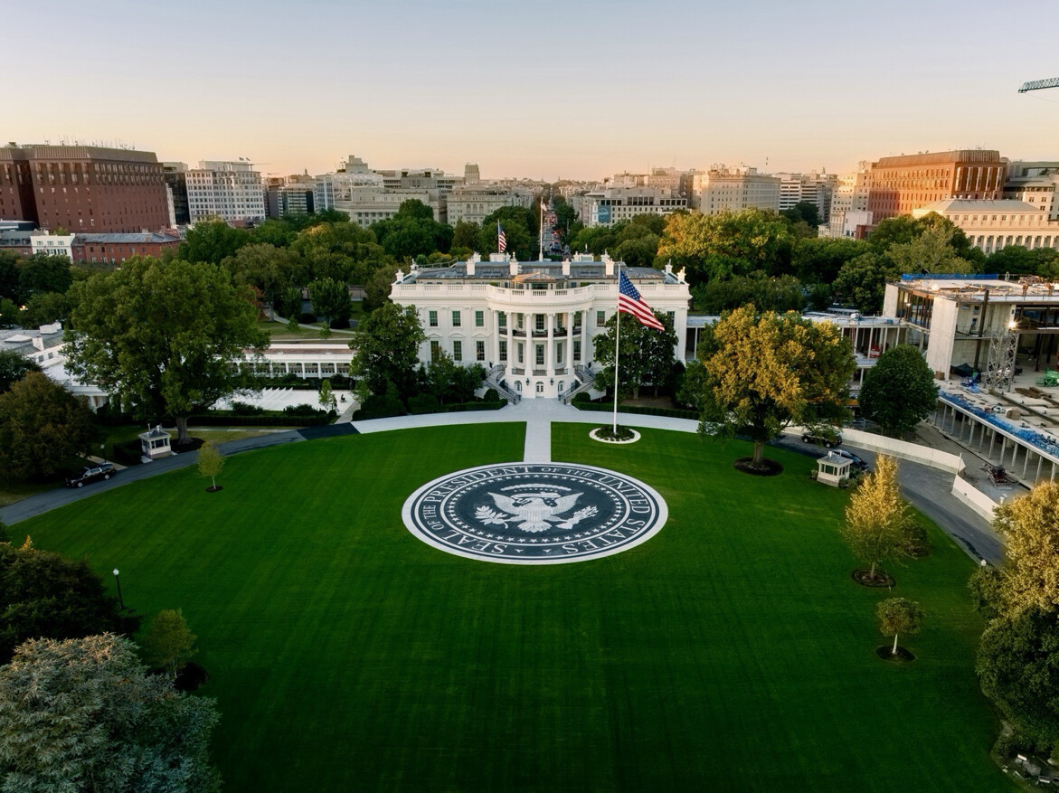 Aerial view of the White House, featuring a prominent circular emblem on the lawn with the text "Seal of the President of the United States." The building is surrounded by trees and flanked by other structures and construction. An American flag is flying in front of the White House.