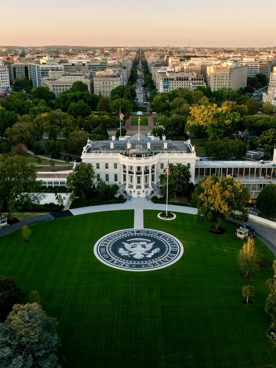 Aerial view of the White House, showcasing its neoclassical architecture and manicured lawn with the Seal of the President of the United States displayed prominently. The image captures the surrounding landscape, including trees, buildings, and streets extending into the distance. Two American flags are visible, one on the roof and another near the seal.