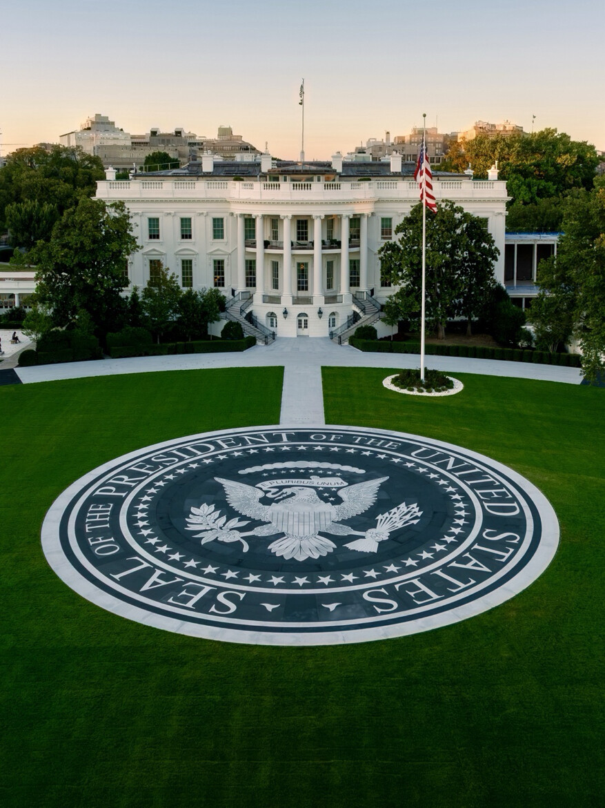 Aerial view of the White House with a prominent circular seal in the foreground on the lawn that reads "Seal of the President of the United States." The seal features an eagle holding arrows and an olive branch, with a shield on its chest and a banner in its beak that reads "E Pluribus Unum." The American flag is visible on a flagpole near the building, surrounded by trees and pathways.
