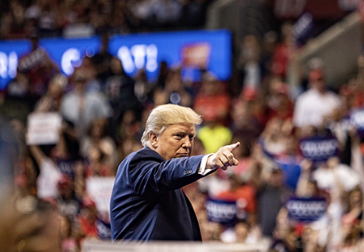 A man with blonde hair and a dark suit is pointing forward with his right hand. He stands in a crowded indoor arena filled with people holding signs. There are blurred blue and red signs in the background, suggesting a political rally or public event.