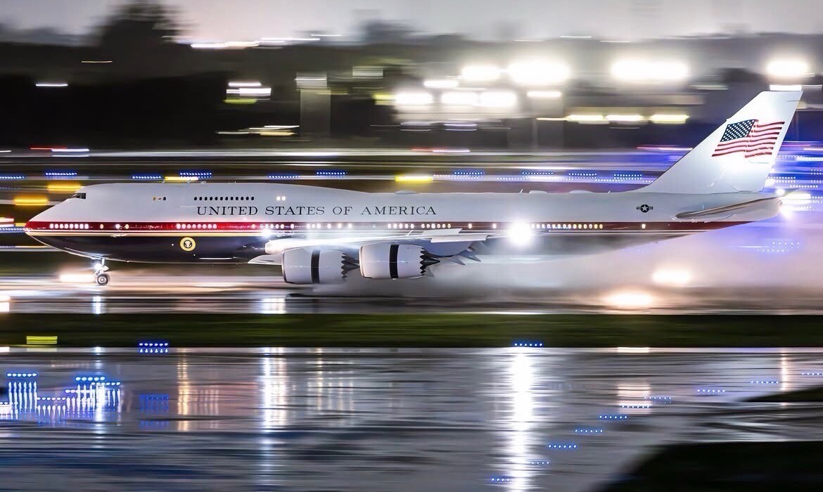 A large aircraft is on a runway at night, with lights reflecting off wet surfaces. The airplane bears the text “UNITED STATES OF AMERICA” and displays an American flag on its tail. The scene is slightly blurred, indicating motion.
