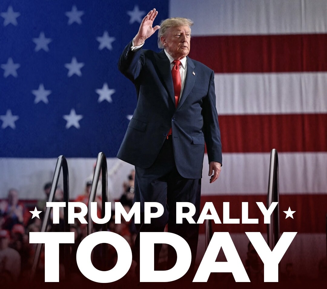 A man in a suit stands on a stage in front of a large American flag backdrop. He is raising his hand in a waving gesture. In the foreground, large text reads "TRUMP RALLY TODAY." The scene is set with a group of people in the background, some of whom appear to be watching the man.