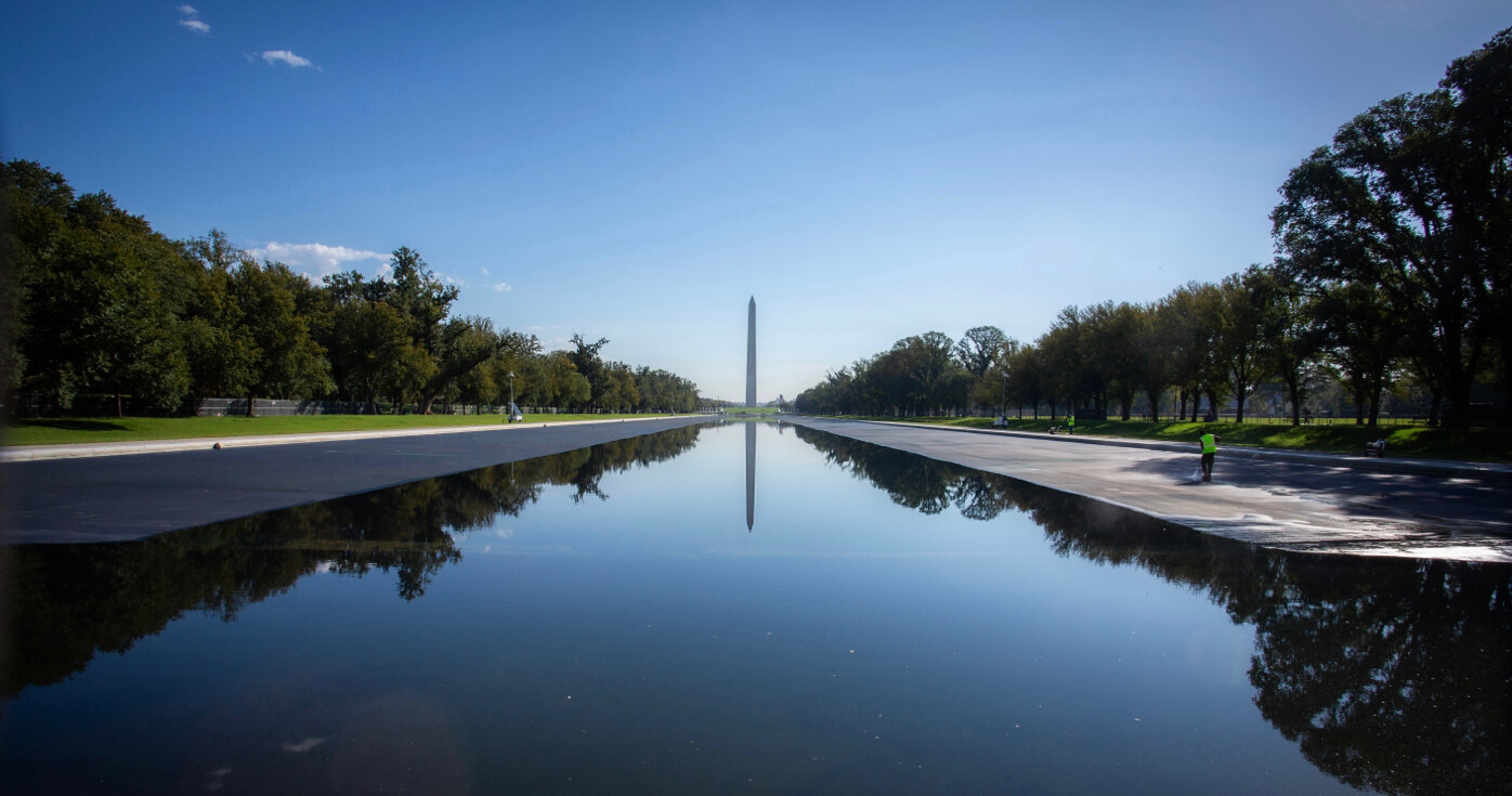 The image shows a clear day view of the Washington Monument, reflected in the Lincoln Memorial Reflecting Pool. The scene is framed by trees on both sides and a few people can be seen near the pool, one wearing a neon safety vest. The sky is blue with scattered clouds.