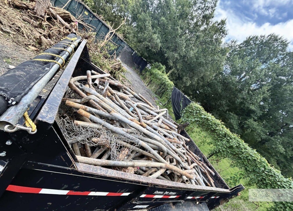 A dump truck is filled with a large quantity of metal pipes and chain-link fencing. The truck is positioned on a gravel area surrounded by vegetation, including trees and bushes. A black mesh tarp is partially covering the contents. There is signage on the side of the truck reading "Caution: Not for hire." The sky is partly cloudy.