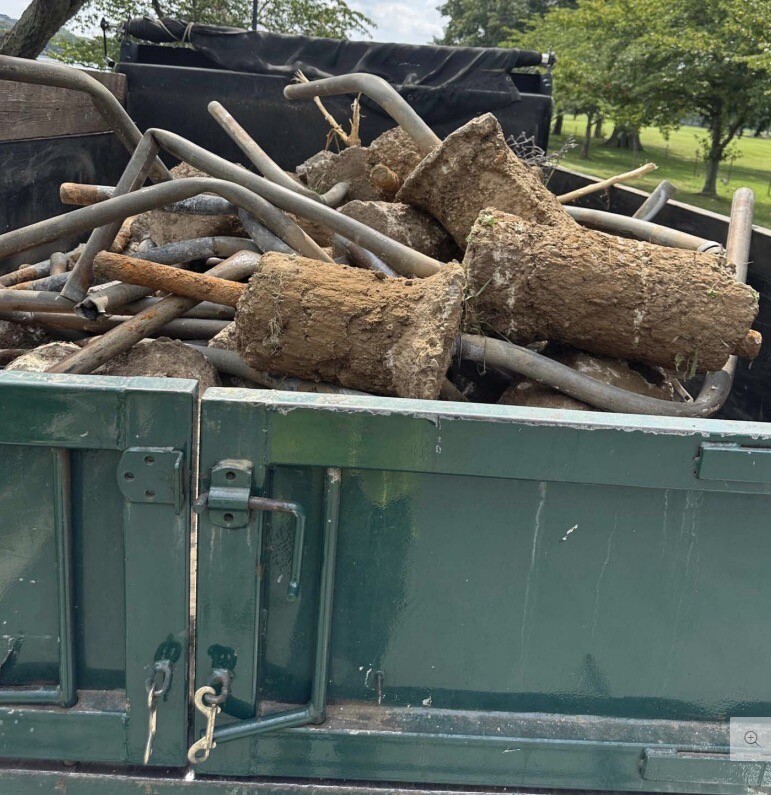 A green truck bed filled with metal pipes and concrete structures, some covered in dirt and grass. The truck bed has a latch and a chain visible on the back gate. Trees and grass are seen in the background.