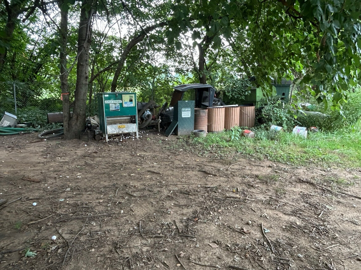 A wooded area with a chain-link fence in the background. There is a green metal gate with a sign that reads "GATE/START" and another sign that says "SWING GATE." Below, there is a smaller sign with "aution" which appears to be part of a caution notice. Next to the gate, there are several wooden crates and scattered barrels on the grassy ground. The scene is outdoors with visible trees and foliage.
