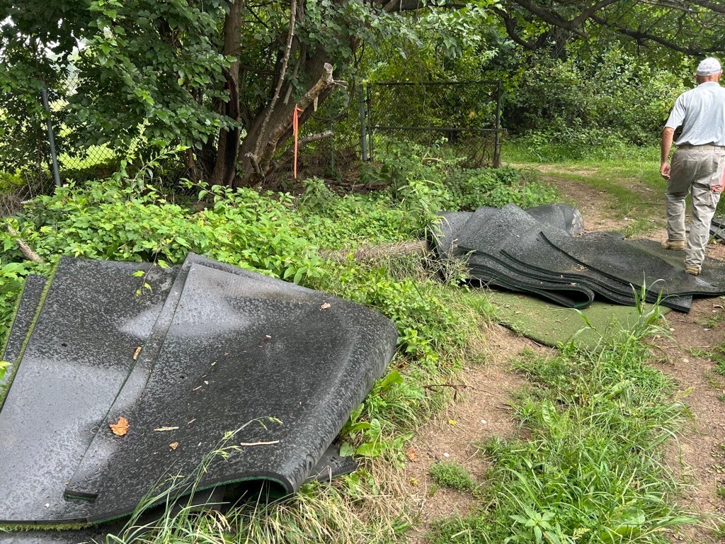 Piles of black mats or rubber sheets are stacked on the ground in a wooded outdoor area, surrounded by green foliage and grass. A person wearing a light gray shirt and beige pants walks away on a dirt path leading into the trees. There is also a metal fence and a large tree nearby.