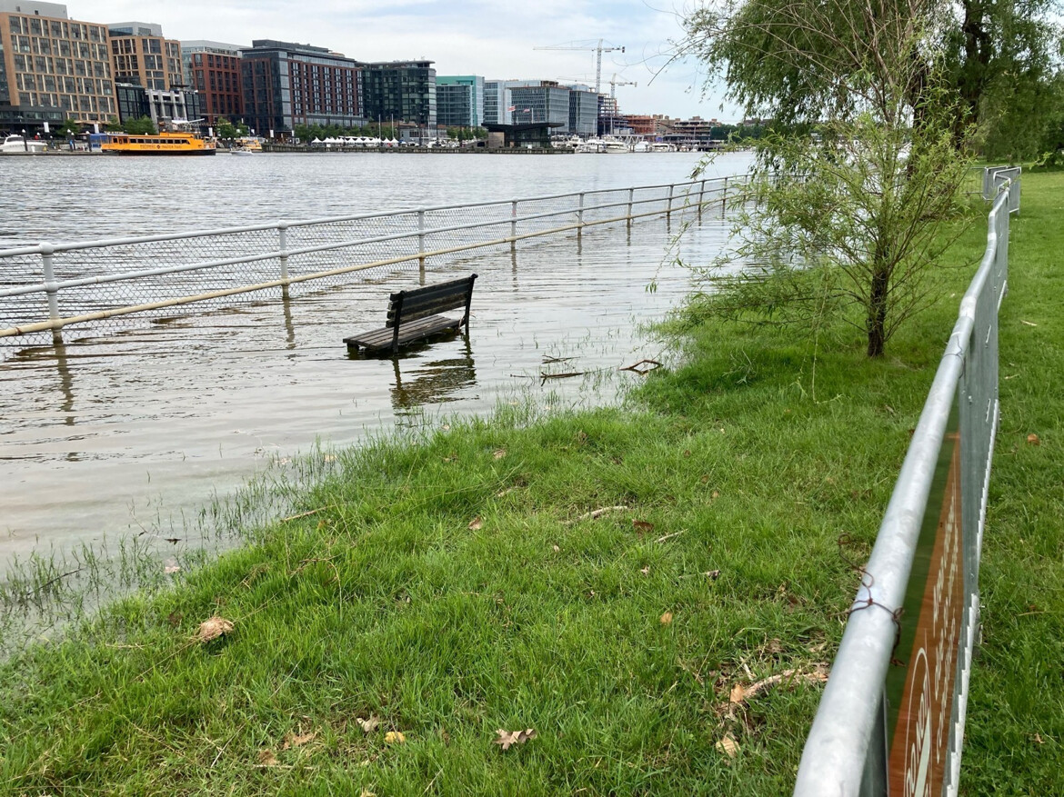 A flooded riverside area with a park bench partially submerged in water. A metal railing runs parallel to the water, leading to a grassy area with a tree. Across the river, modern buildings are visible, along with a yellow boat and construction cranes, indicating an urban setting.