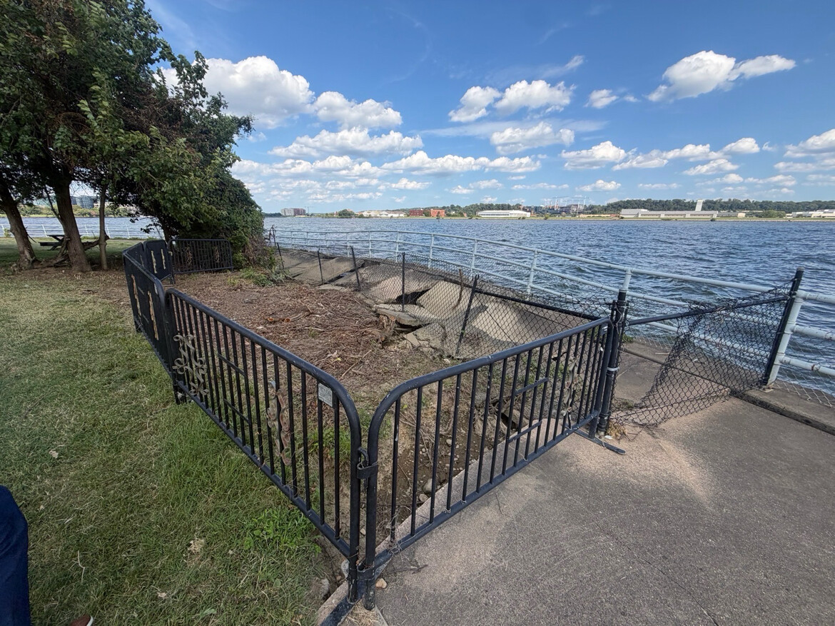 A waterfront scene with a damaged metal fence, part of which is bent and detached, surrounded by grass and a few trees on the left. The view includes a body of water on the right, with a partly cloudy sky and some industrial buildings in the distance.