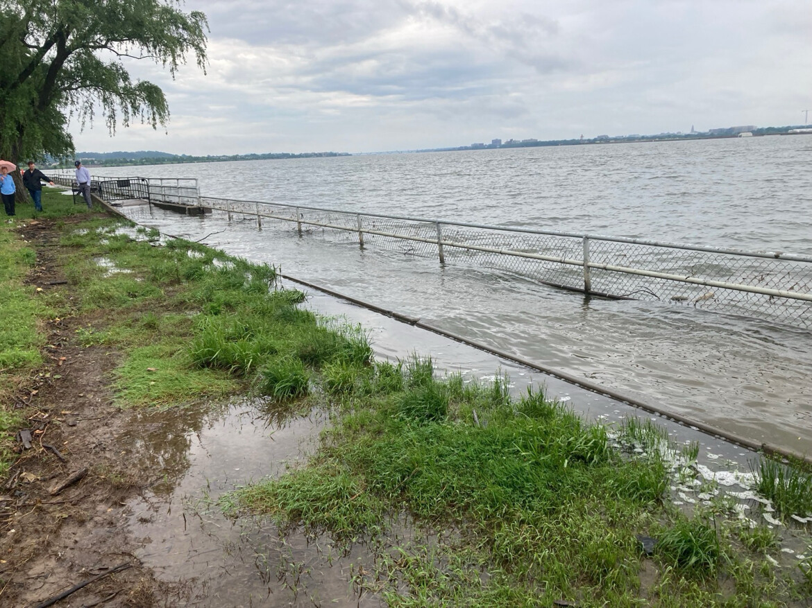 A flooded riverside area shows a chain-link fence partially submerged in water, with the river levels rising over the walkway. Three people stand on the grassy area nearby, observing the situation. The sky is overcast, suggesting recent or ongoing rain.