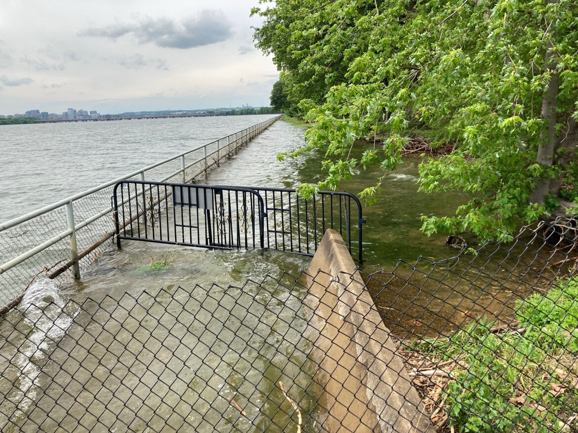 A flooded pathway with a metal fence gate partially submerged in water. The path runs parallel to a large body of water, with a railing on one side and trees on the other. A city skyline is visible in the distance under a cloudy sky. The scene indicates high water levels encroaching on the pathway.