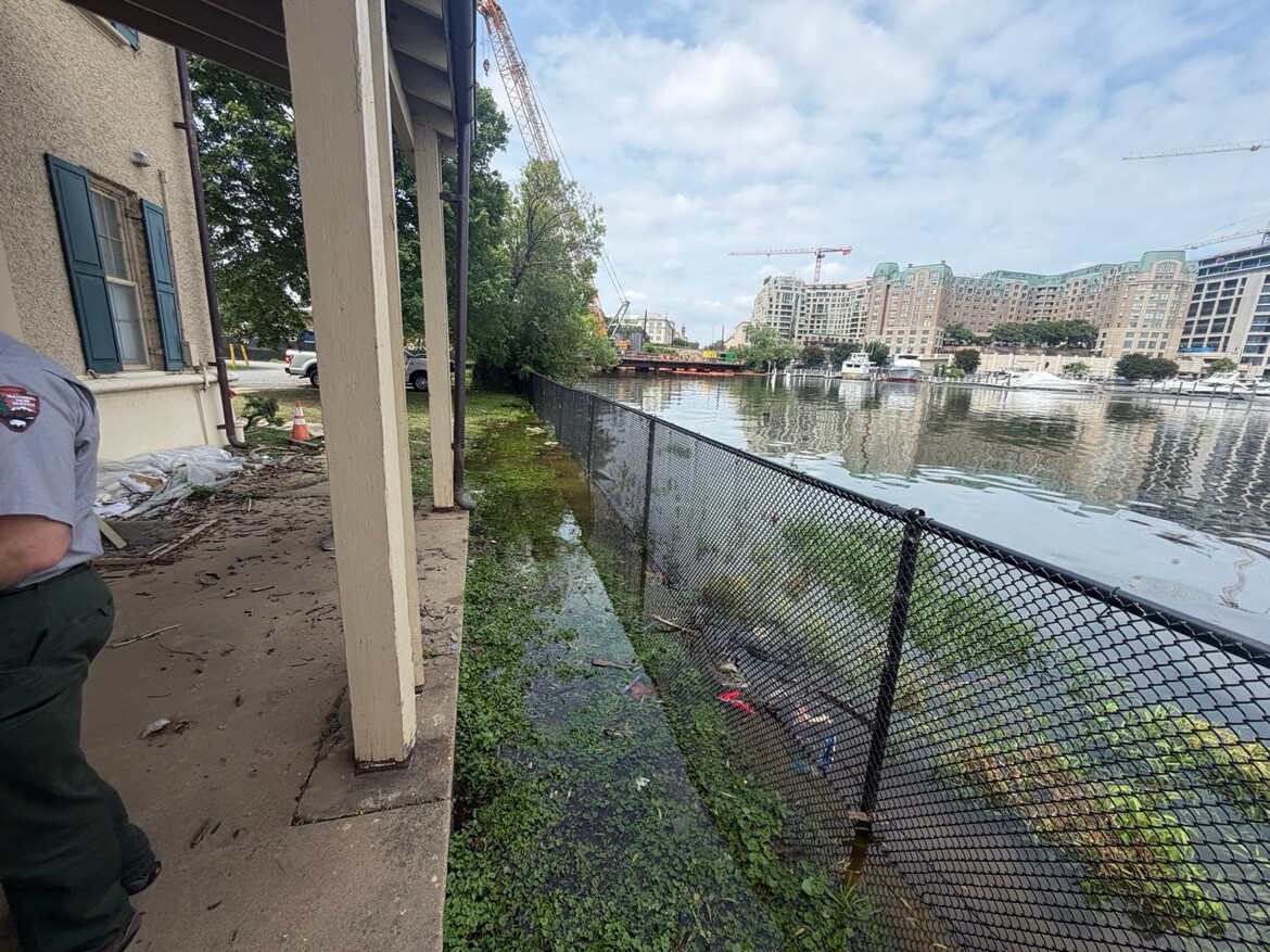 A partially flooded walkway next to a building is shown, with water spilling over into the path. A chain-link fence runs along the water's edge, separating the walkway from a body of water on the right. Several buildings and cranes are visible across the water, suggesting nearby construction. A person in a uniform stands on the left side of the image, partially out of view.