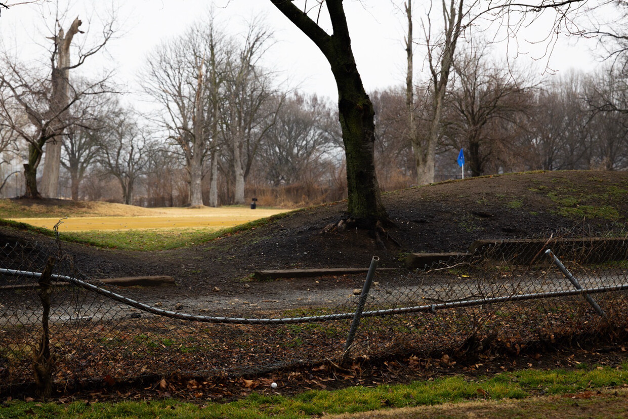 A park scene with bare trees and grassy areas. A chain-link fence in the foreground is bent and damaged. In the background, there is a blue flag on a pole, possibly marking a golf course or field area. The ground is a mix of grass and bare soil, and the atmosphere appears overcast.