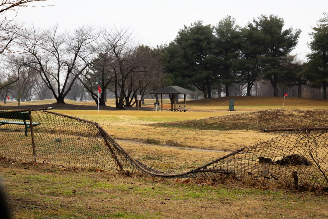 A golf course with a damaged chain-link fence in the foreground, partially bent and slumped towards the ground. In the background, there are trees with bare branches, a small pavilion with benches, and several red flags marking different holes. The landscape has a mix of green and brown patches, suggesting an early spring or late winter setting.