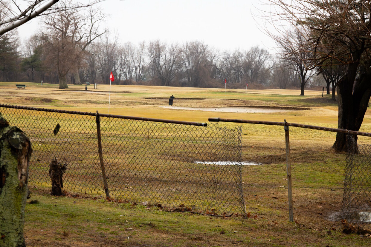 A golf course with several red flags marking holes is visible beyond a chain-link fence. The grass appears patchy and dormant, common in colder seasons. Trees without leaves surround the area, indicating a winter scene. A bench and trash can are visible in the background.