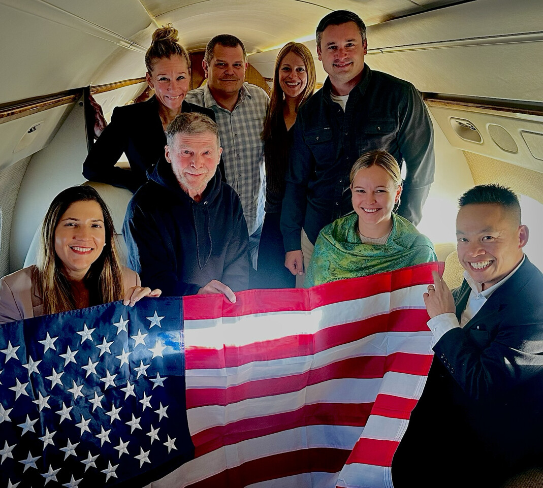 A group of eight people are inside a private jet, holding an American flag. They are arranged closely together, smiling at the camera. The interior of the jet is visible, with windows and seats. The people are dressed in casual and business attire.