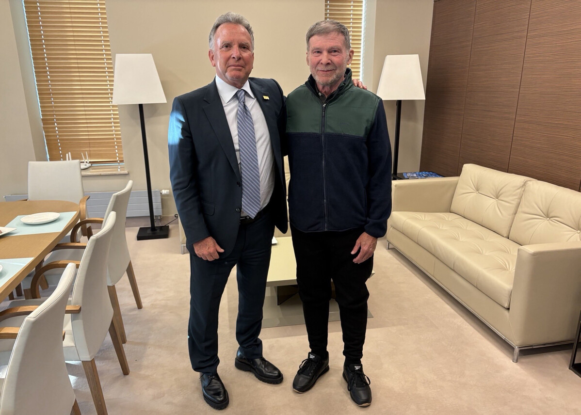 Two men stand together in a well-furnished office, with one man wearing a formal suit and tie while the other wears a casual jacket and pants. The room features a table with white chairs, a white couch, lamps, and a window with blinds. Both men are smiling and posing for the photograph.