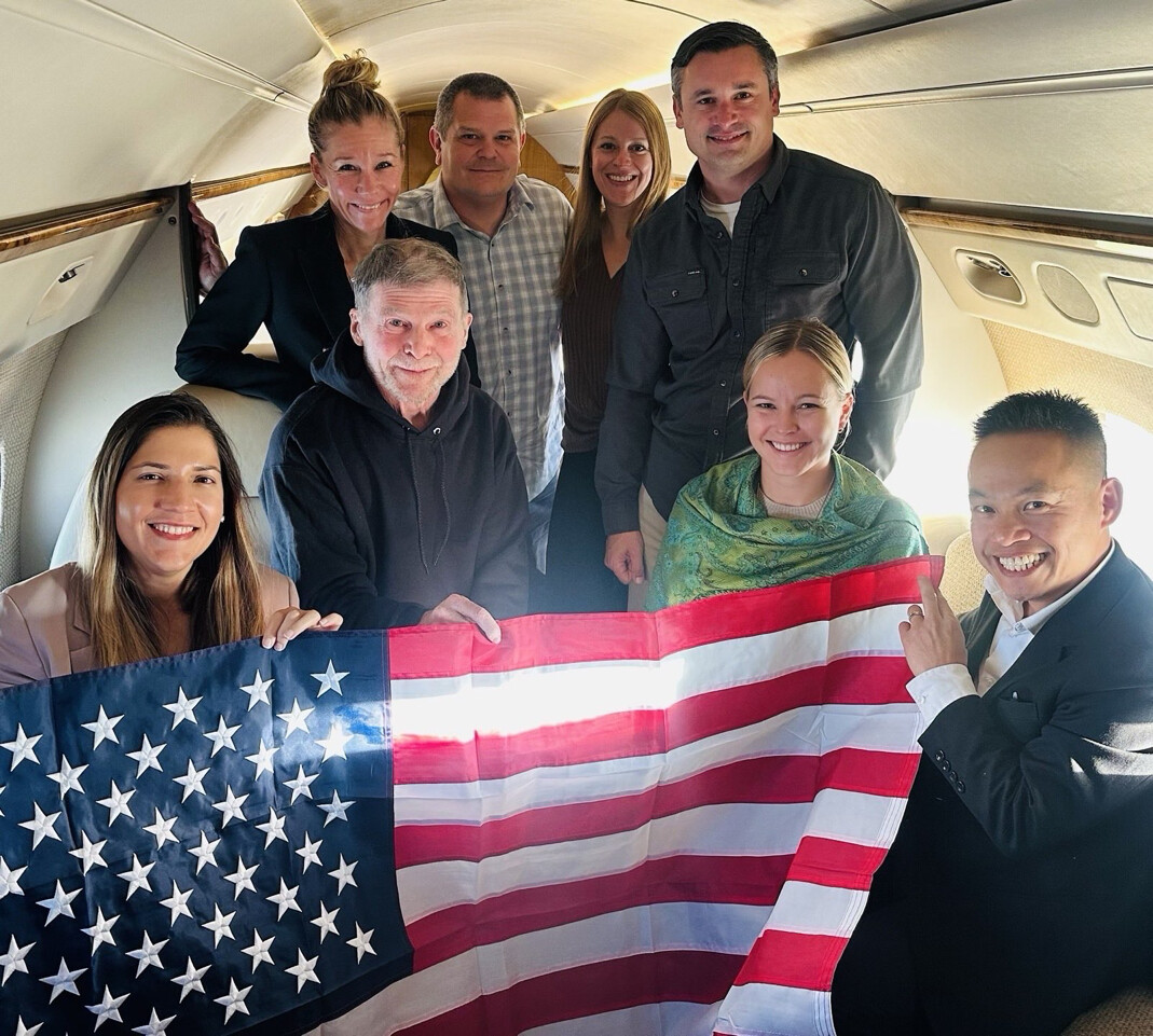 A group of eight people is posed inside an airplane cabin, holding a large American flag. The individuals are smiling and dressed in a mix of casual and formal attire. The interior of the airplane appears luxurious, with visible seats and lighting.