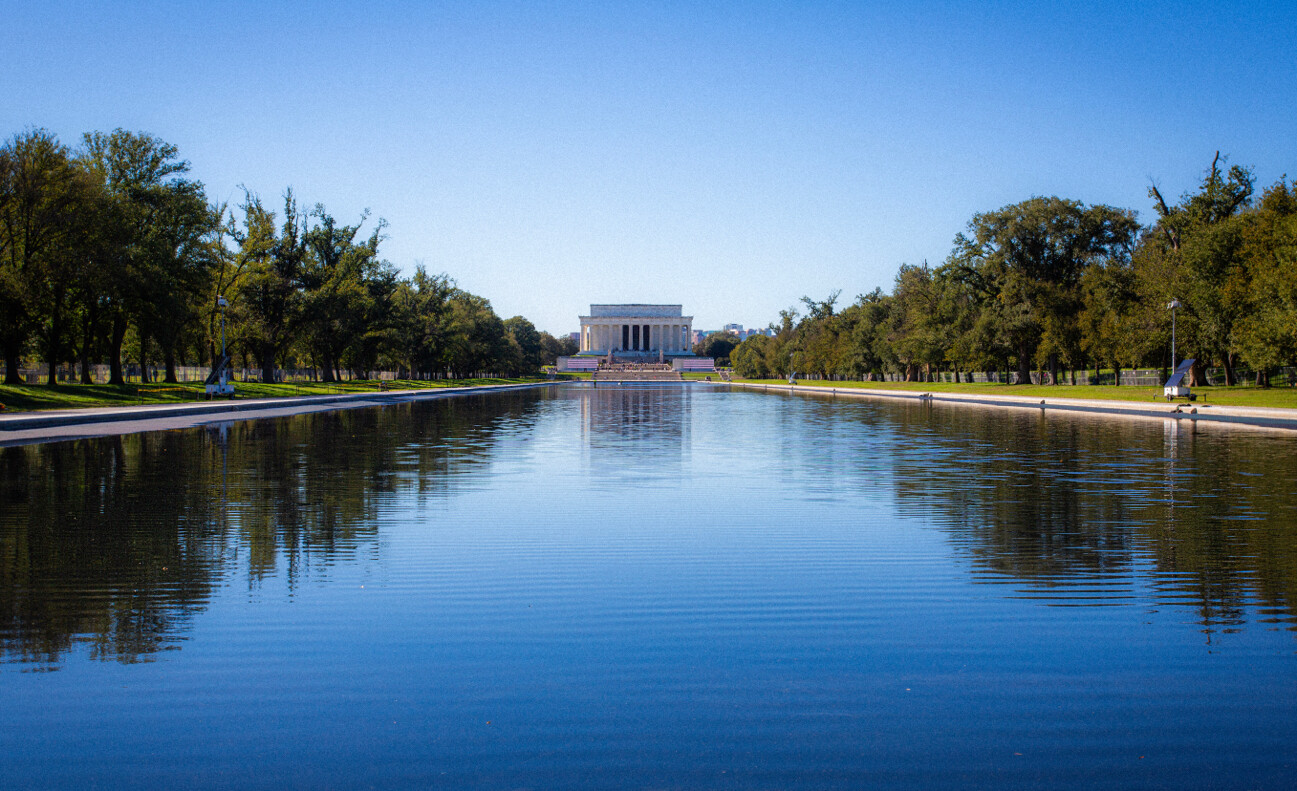 A wide-angle view of the Lincoln Memorial in Washington, D.C., seen from the opposite end of the Reflecting Pool. The pool is flanked by trees and walkways on both sides, under a clear blue sky. The scene is symmetrical, with the monument centered in the background.