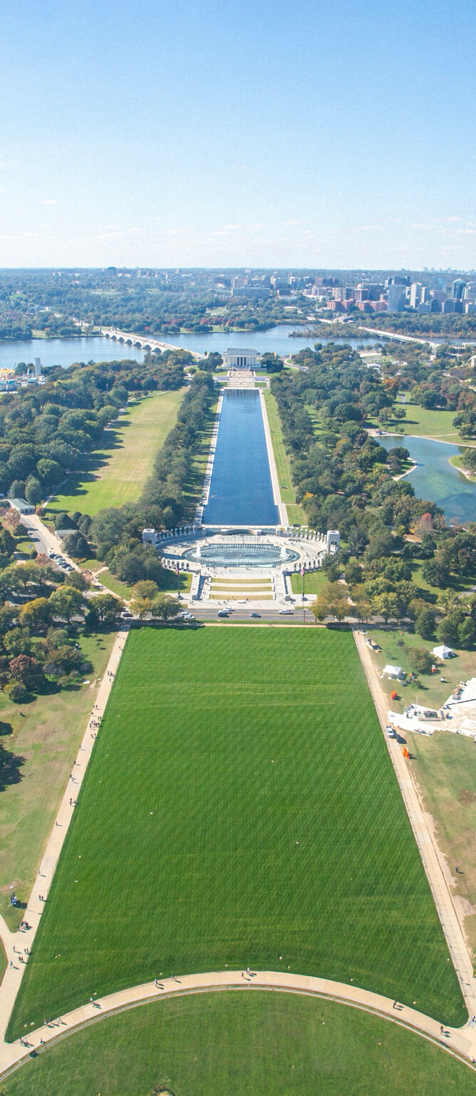Aerial view of the National Mall in Washington, D.C., featuring the Lincoln Memorial, Reflecting Pool, and World War II Memorial. The image shows a large expanse of green lawns and pathways, with the Potomac River and Arlington Memorial Bridge visible in the background. The surrounding area is filled with trees and landscaped parks, leading to the city skyline.